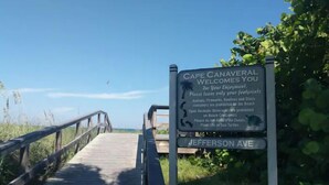 A wooden boardwalk leading to a beach with a sign that reads "Cape Canaveral Welcomes You" and mentions Jefferson Ave.