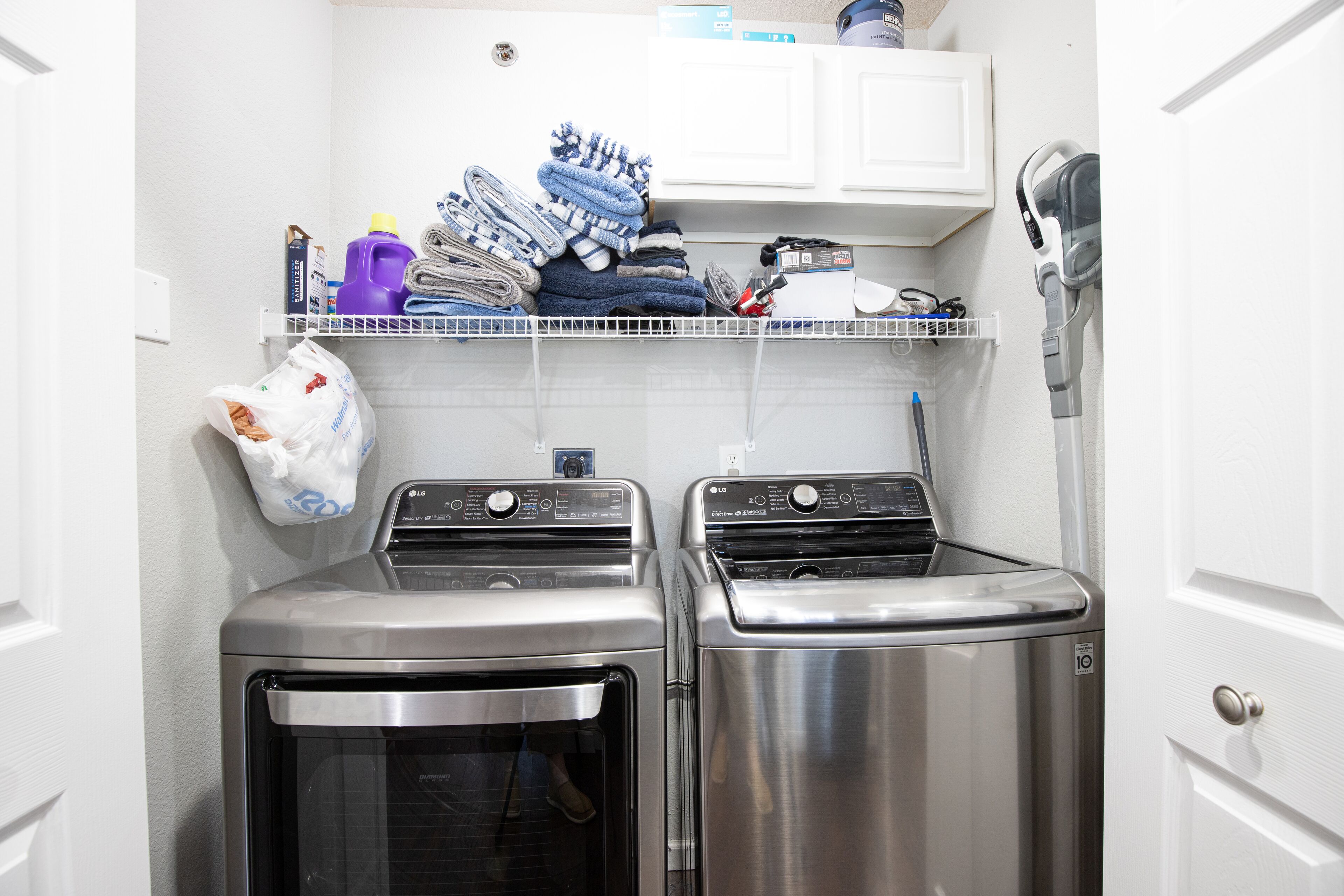 A laundry room with two stainless steel dryers, a shelf with folded towels and laundry supplies, and a bag of laundry on the wall.