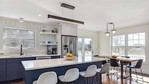 A modern kitchen with a central island, bar stools, and a dining area with a table and chairs. - Amazing harbor/ocean views of Pepperrell Cove (Kittery)
