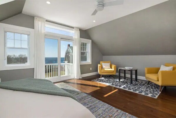 Une chambre avec un grand lit, un balcon avec vue, deux fauteuils jaunes et un tapis à motifs.