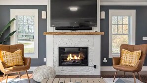 A living room with a fireplace, two armchairs, a TV mounted above the fireplace, and a large window on the left. - Amazing harbor/ocean views of Pepperrell Cove (Kittery)