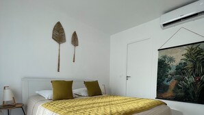 A bedroom with a bed, two pillows, a nightstand, a wall-mounted air conditioner, and a painting.