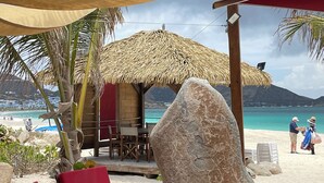 A beachside lounge with red cushioned chairs and a wooden table, a thatched-roof structure, and a large rock in the foreground.