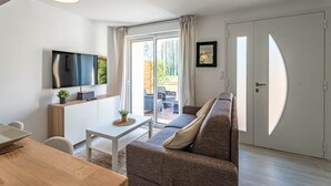 A modern living room with a sofa, a TV, a dining table, and a sliding glass door leading to an outdoor area. - La Grange du Noé: L'Odon (Caen)