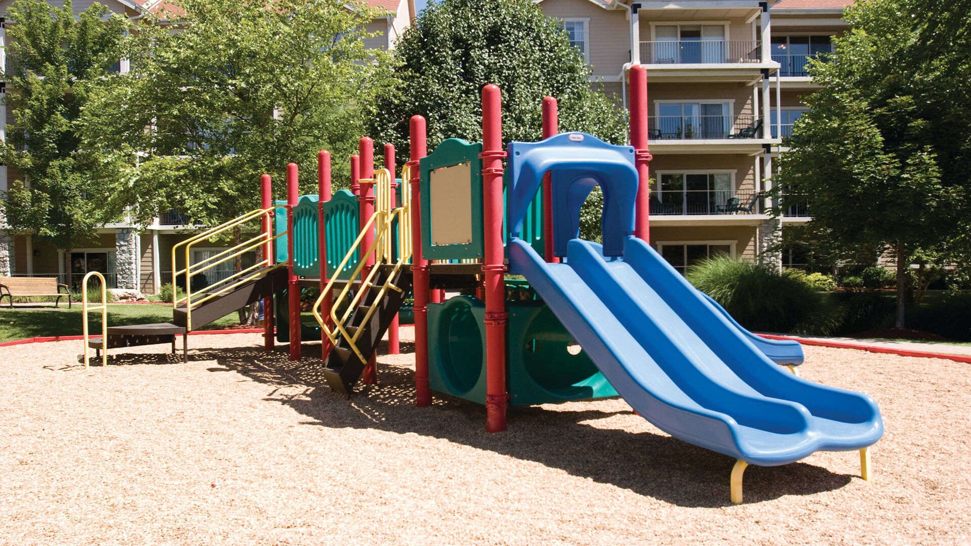 A playground with a slide and climbing structures, surrounded by a building with balconies and a tree.