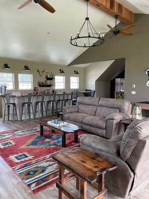 A spacious living room with a large gray sofa, a wooden coffee table, and a colorful rug. There is a kitchen area with bar stools and a chandelier. - Sunset Serenade in the Country (Eighty Four)