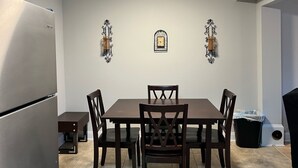 A dining area with a table and chairs, a clock on the wall, and a trash can.