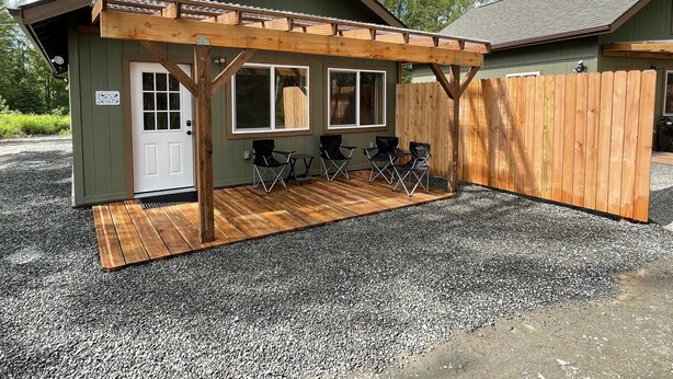 Una cabaña pequeña con terraza de madera, dos sillas plegables y un porche cubierto.