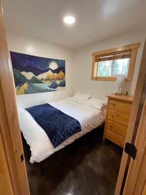 A bedroom with a bed, a dresser, a night sky painting, and a window with blinds.