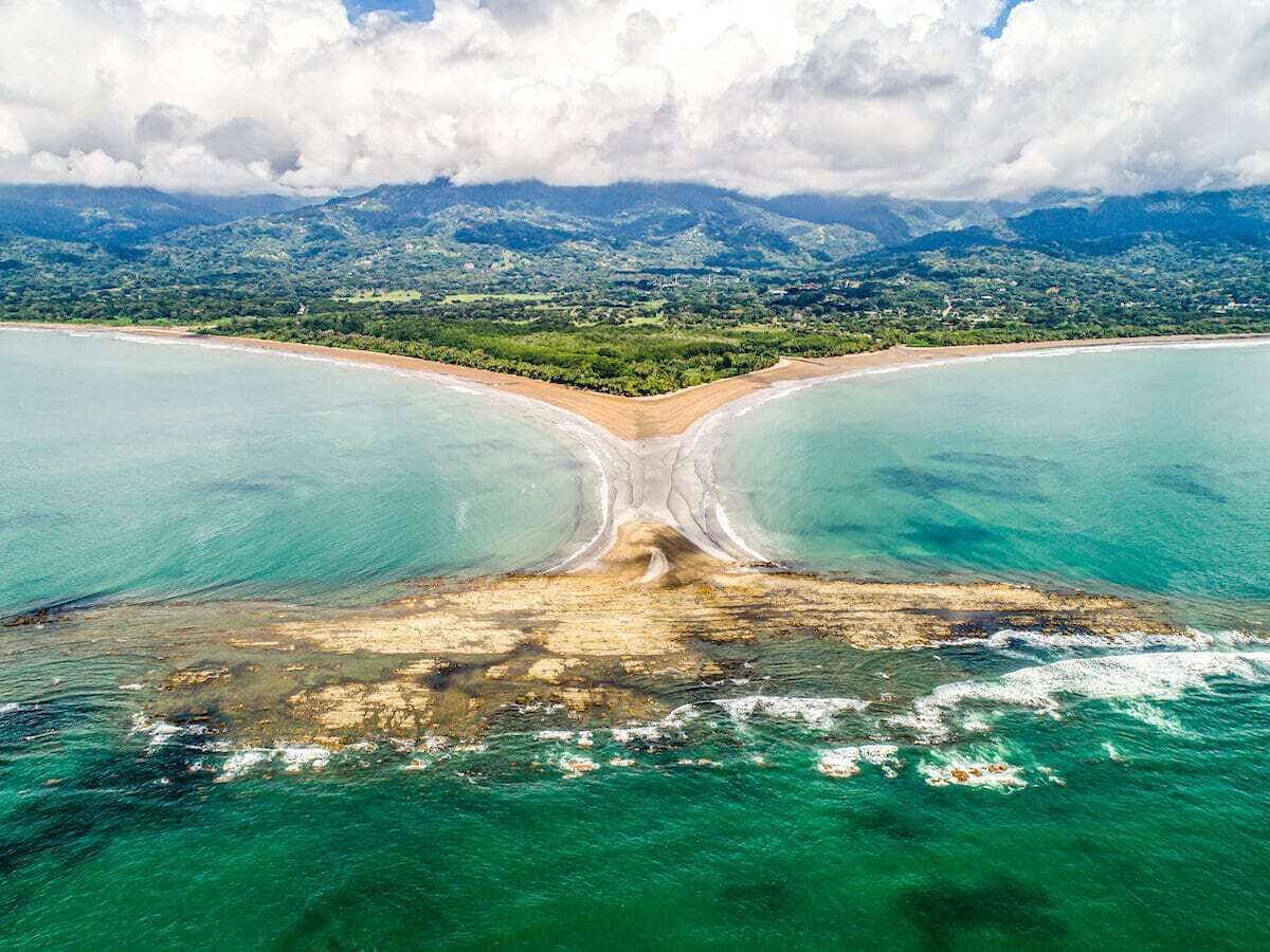 Une vue aérienne d’une plage où l’on distingue une séparation nette dans le sable, entourée d’eaux turquoise et dominée par un paysage montagneux en arrière-plan.