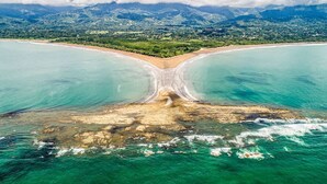 Eine Vogelperspektive zeigt einen Strand mit einer deutlichen Sandspalte, umgeben von türkisfarbenem Wasser und einem Bergpanorama.