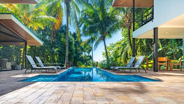 Ein Schwimmbecken mit zwei Liegestühlen an jeder Seite, umgeben von Palmen und einer gefliesten Terrasse.