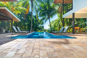 Une piscine bordée de deux chaises longues de chaque côté, entourée de palmiers et d’une terrasse carrelée.
