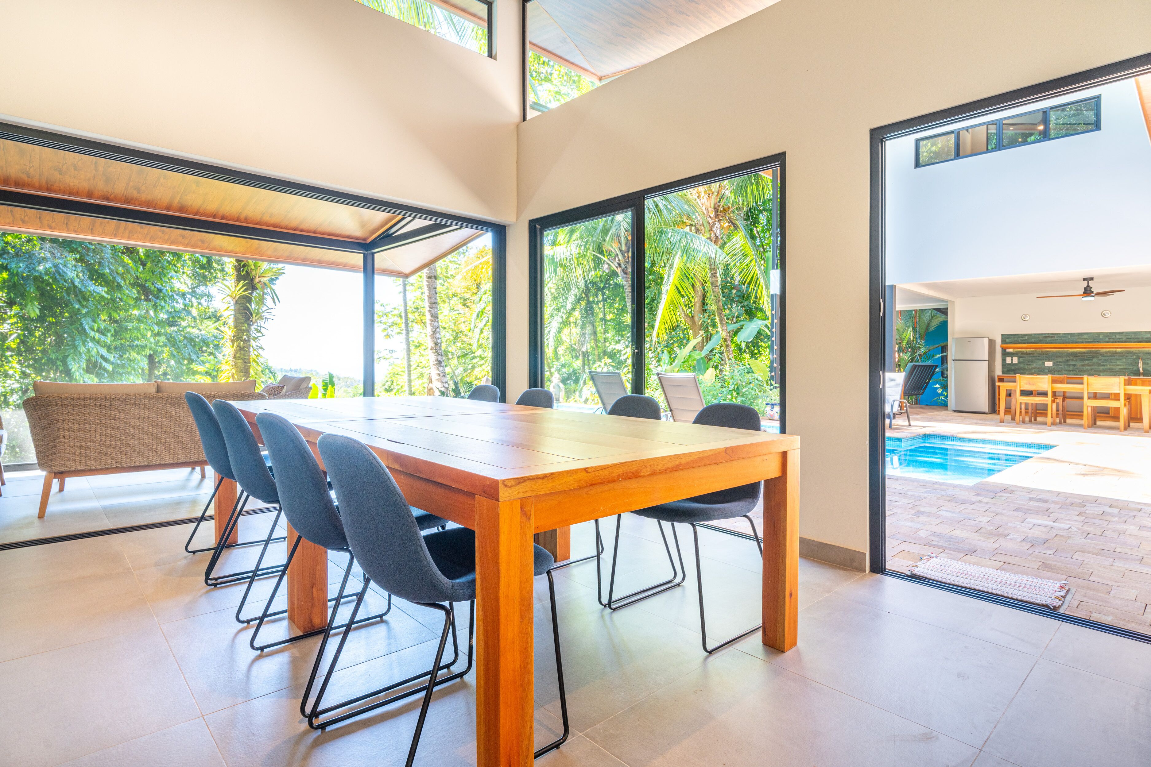 Un comedor moderno con una amplia mesa de madera y sillas, ventanas grandes con vistas a la vegetación, y una piscina visible a través de puertas de cristal.
