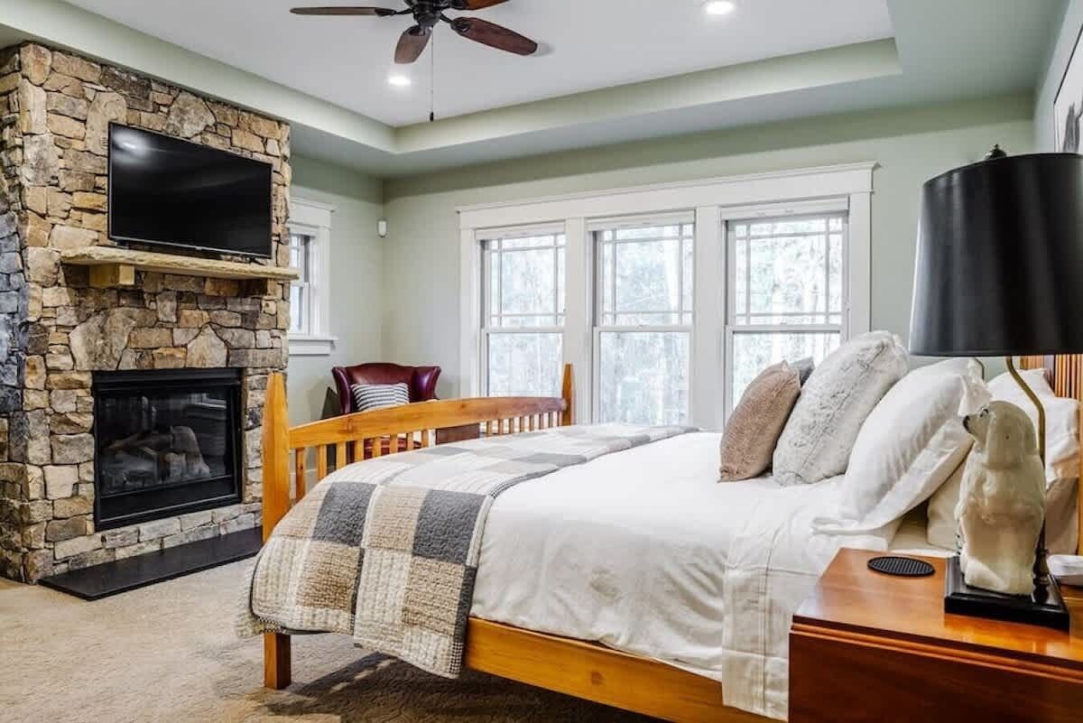 Une chambre avec une cheminée en pierre, un lit recouvert d’une courtepointe, une tête de lit en bois, un ventilateur de plafond et une fenêtre avec des rideaux blancs.