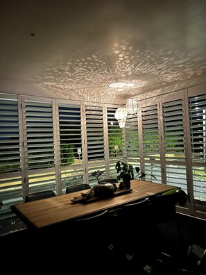 A room with a wooden table, a hanging light fixture, and a view through large windows with blinds. - Luxury At The Lookout - Park and Beach Across The Road! (Ocean Grove)