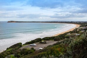 A coastal landscape with a beach, parked cars, and a distant cliff. - Luxury At The Lookout - Park and Beach Across The Road! (Ocean Grove)