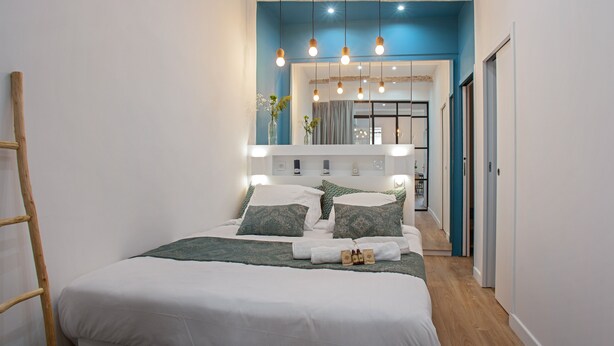 A modern bedroom with a large bed, bedside lamps, and a view into the living area. - Loft L'Escale - 3 étoiles - Welkeys (La Ciotat)