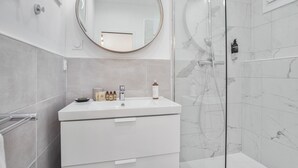 A modern bathroom with a white vanity, a round mirror, and a glass-enclosed shower area. - Loft L'Escale - 3 étoiles - Welkeys (La Ciotat)