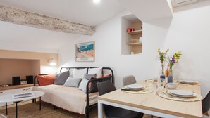 A modern, well-lit room with a bed, a dining table set for two, and a small shelf with decorative items. - Suite Mugel - 3 étoiles - Welkeys (La Ciotat)