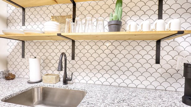 Cozinha com bancada de granito, pia de aço inoxidável e backsplash com padrão em forma de concha. Duas prateleiras de madeira acima da bancada exibem copos e um vaso de planta.