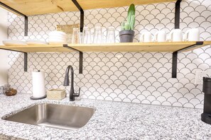 A kitchen with a granite countertop, stainless steel sink, and a backsplash with a scalloped pattern. There are two wooden shelves above the countertop, holding glassware and a potted plant. - The Bohemian- 1/2 mi from the Covington Square (Covington)