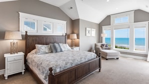 A bedroom with a large bed, two bedside tables, a chair, and a view of the ocean through large windows. - Coastal LunaSea: Oceanfront, Pool, Elevator OBX (Corolla)