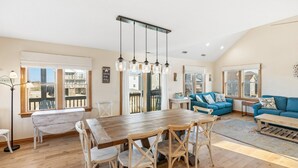 A spacious living area with a wooden dining table, blue sofas, and large windows offering views of the outside. - Cedar Sandcastle*semi-oceanfront*Hot Tub*Foosball (Nags Head)