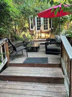 A wooden deck with outdoor furniture, a red umbrella, and a wooden house in the background. - Cozy Creekside Cabin in Santa Cruz Mountains (Soquel)