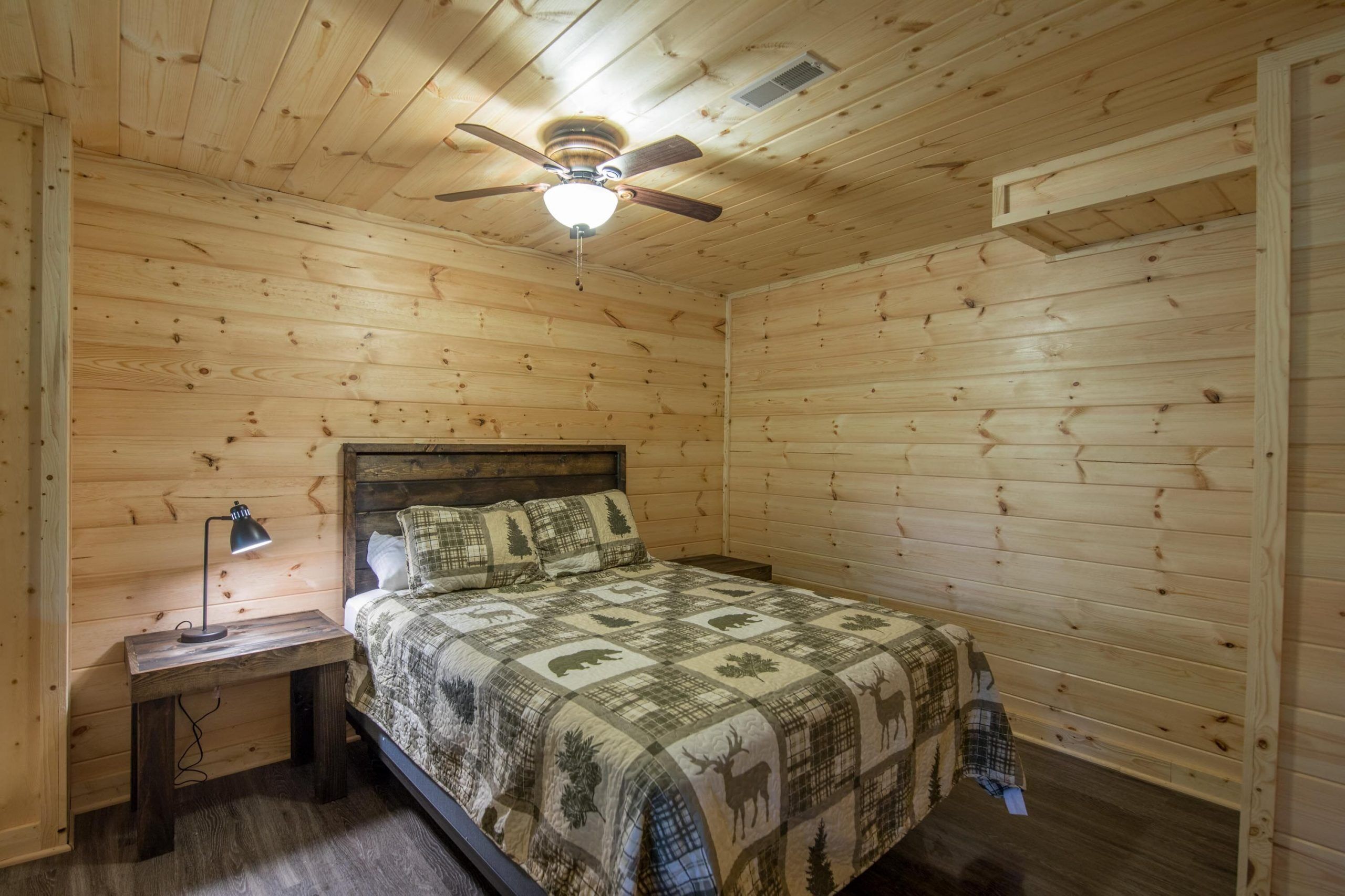 A wooden-paneled bedroom with a bed, a nightstand, a desk lamp, and a ceiling fan.
