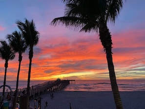 A beach with a wooden pier extending into the ocean, palm trees, and a sunset.