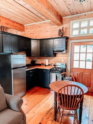 Interior de una cabaña de madera con cocina, que incluye gabinetes negros, un refrigerador de acero inoxidable, una estufa y una mesa de comedor con sillas.