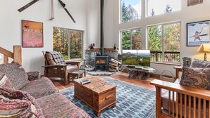A cozy living room with a stone fireplace, wooden furniture, and large windows offering a view of the outdoors.