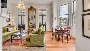Un salon avec un canapé vert, une table basse, un lustre, une table à manger avec des chaises et de grandes fenêtres.