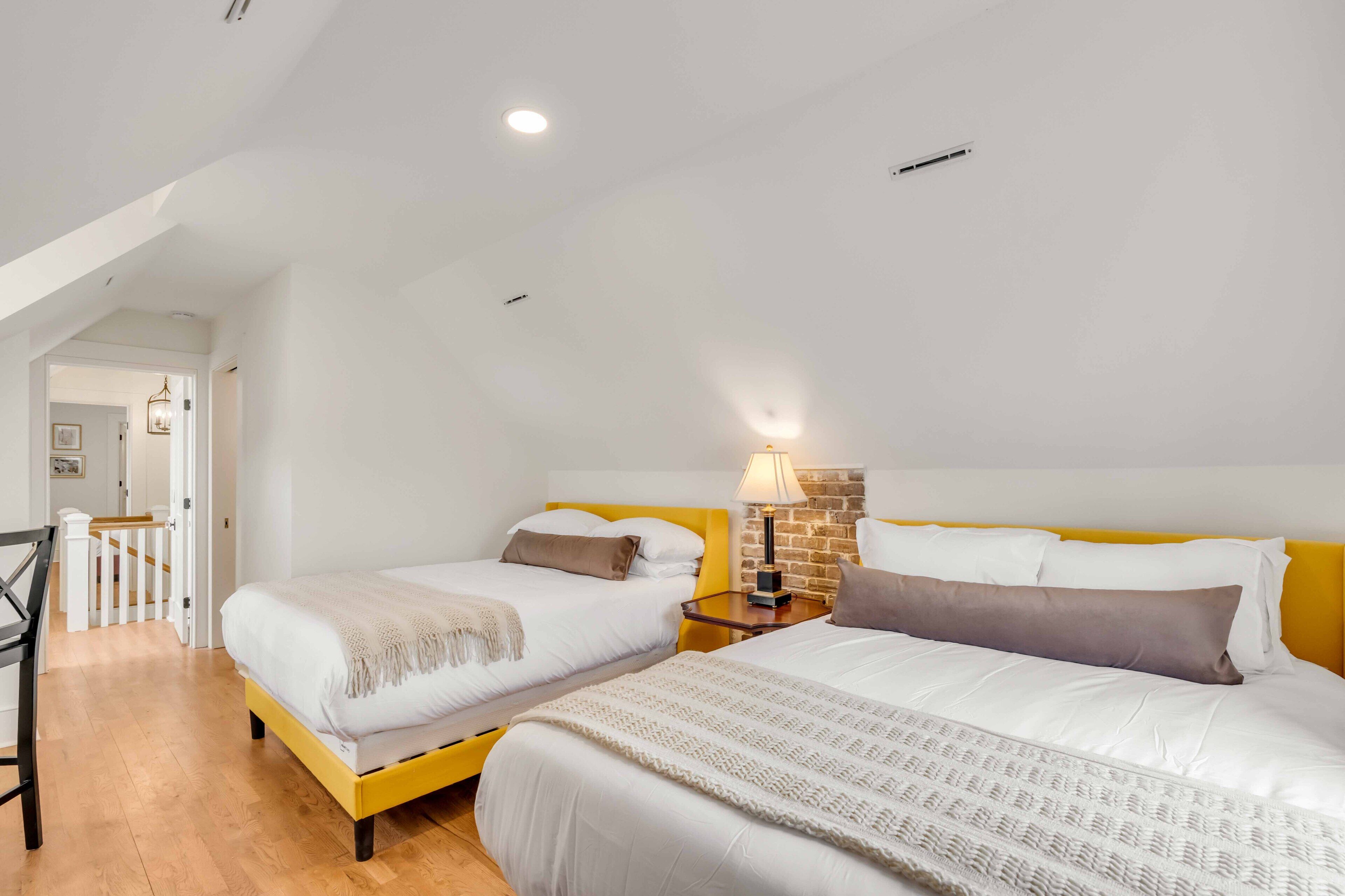 A bedroom with two beds, a wooden floor, a white ceiling, and a small table with a lamp.
