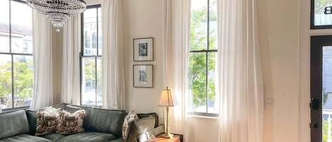 A living room with a green sofa, orange chairs, a coffee table, a chandelier, and large windows with white curtains.