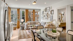 A modern living room with a sofa, coffee table, and dining table. There are curtains, a lamp, and framed pictures on the walls.