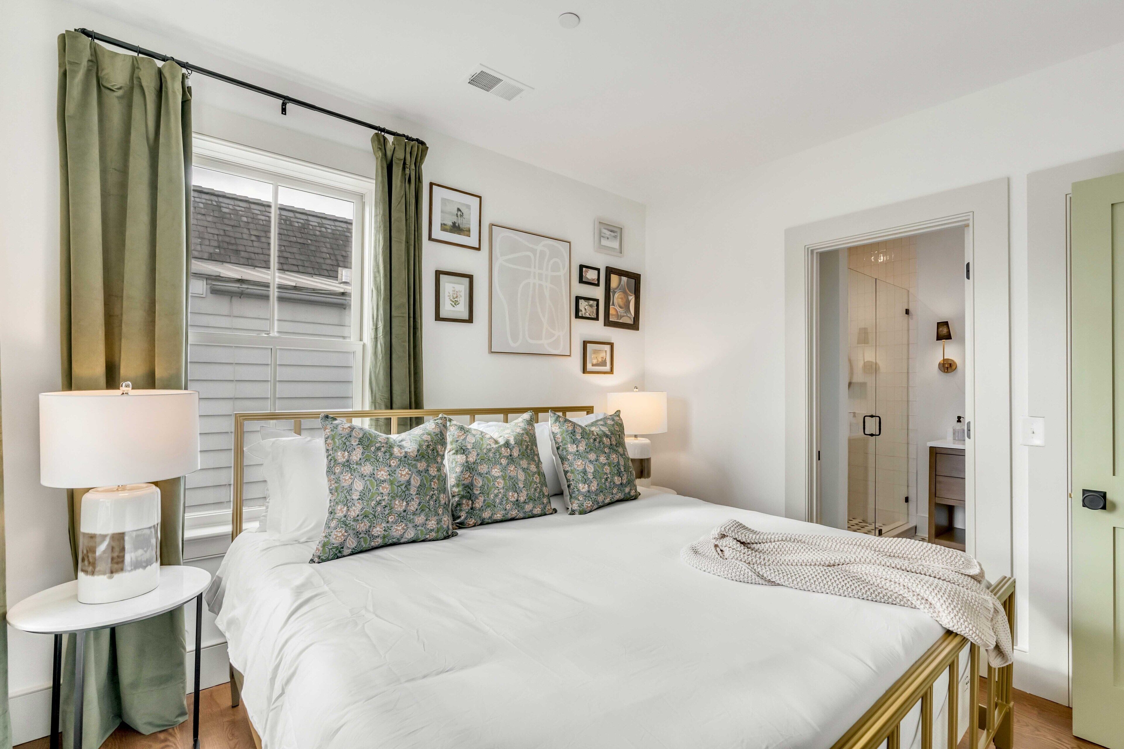 A neatly arranged bedroom with a bed, two bedside lamps, a window with curtains, and a door leading to a bathroom.