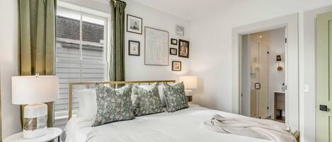 A neatly arranged bedroom with a bed, two bedside lamps, a window with curtains, and a door leading to a bathroom.