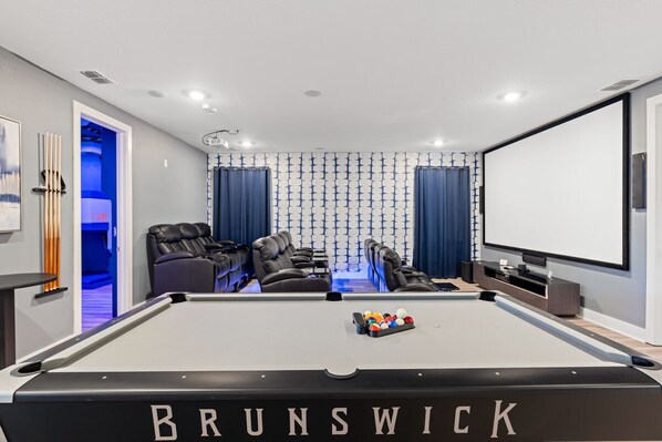 A modern home entertainment room with a pool table, reclining chairs, a projector screen, and a large TV. - Marvelous Muirfield | 10 BR, Ultra Themed Kids Rooms, Movie Theater (Kissimmee)