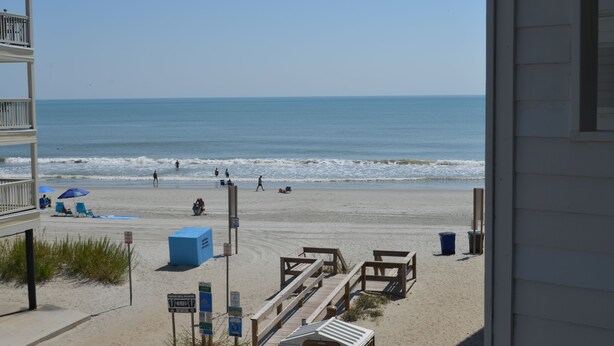 Uitzicht op het strand vanaf een balkon, met zand, enkele mensen, een blauwe parasol en houten constructies.