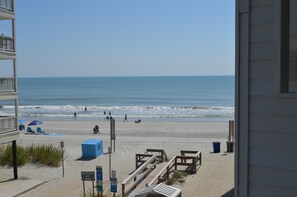 A beach view from a balcony overlooking a sandy beach with a few people, a blue umbrella, and wooden structures.
