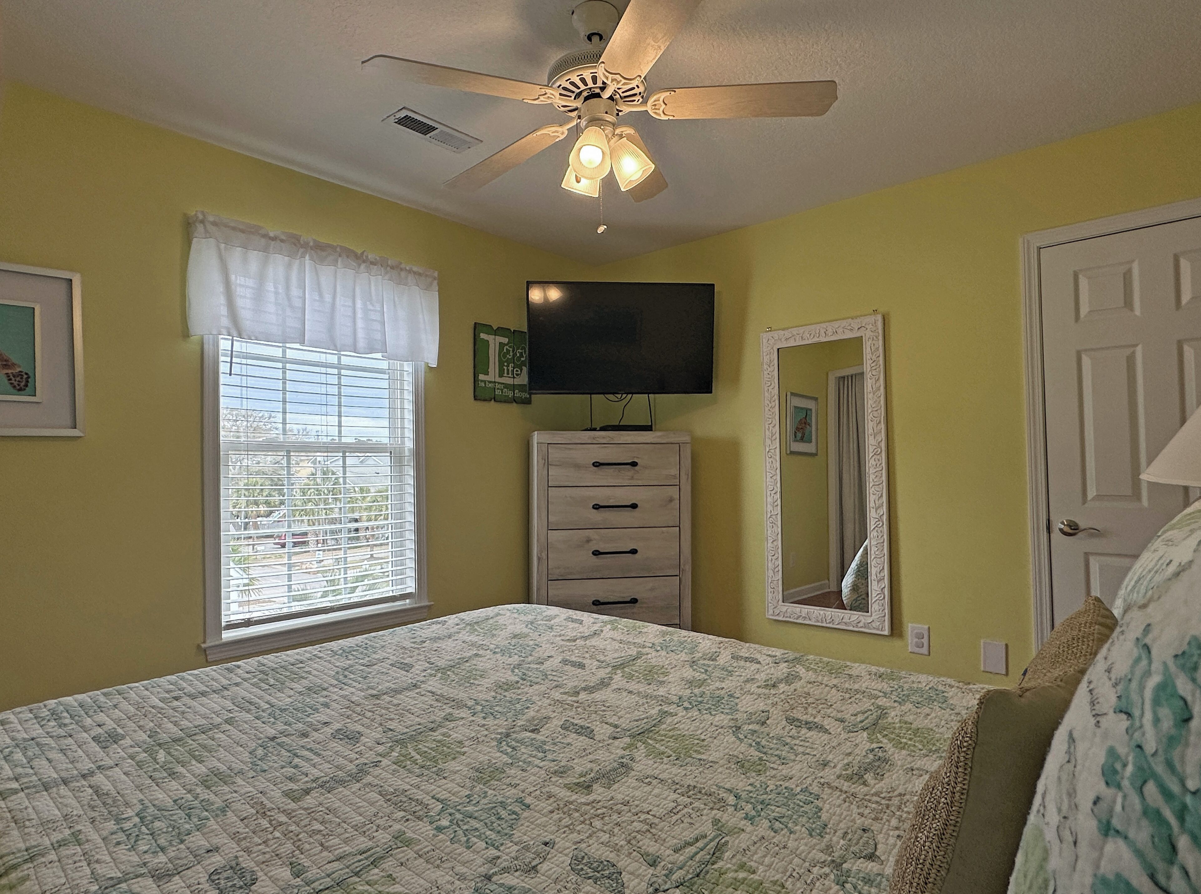 Une chambre à coucher comprenant un lit, un ventilateur de plafond, une commode, un téléviseur et une fenêtre avec des stores.