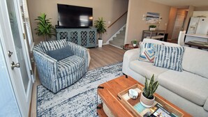 Ein Wohnzimmer mit einer Couch, zwei Sesseln, einem Couchtisch, einem Fernseher und einer Treppe, die in einen anderen Raum führt.