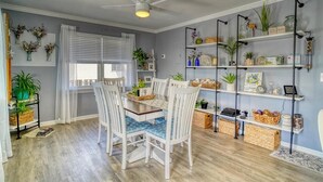 Un comedor con una mesa, sillas y un ventilador de techo. Se observan estanterías con plantas y objetos decorativos en la pared.