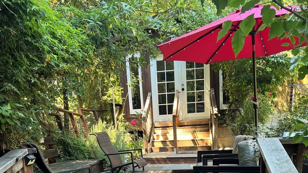 Una terraza de madera con mobiliario exterior, un paraguas rojo y una casa de madera al fondo.