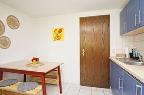 Una cocina pequeña con una mesa de madera, un lavamanos y armarios. Hay una puerta con picaporte y una imagen de tulipanes en la pared.