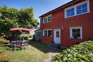 Una casa roja de dos pisos con un pequeño jardín, un patio con sillas y una mesa, y un paraguas rojo.