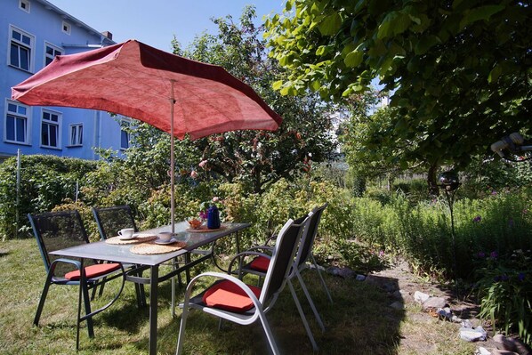 A garden patio with a table and chairs, a red umbrella, and a blue building in the background.