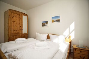 Ein Schlafzimmer mit großem Holzschrank, einem Bett mit weißer Bettwäsche, zwei gerahmten Bildern an der Wand und einem Nachttisch mit Lampe.